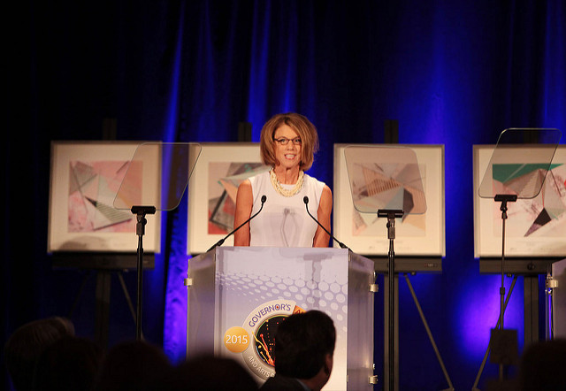 Woman standing at podium giving speech at the Ohio Arts Council 2015 Governor's Awards event. Art pieces on stage behind her.