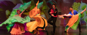 Five dancers in colorful costumes dance exuberantly on stage in front of a botanical backdrop.