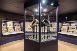 An exhibition room with a central glass case showcasing two large stone artifacts. Surrounding displays feature various archaeological items like pottery and tools, arranged in well-lit cases along the walls. The setting appears modern, designed to highlight ancient cultural artifacts.